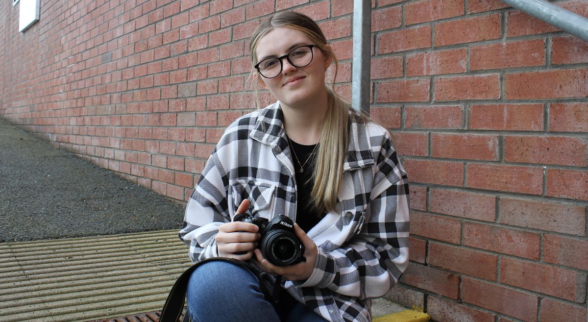 Young woman, Erin O'Neill, with long blond hair, wearing glasses, check shirt and jeans, holding camera and sitting on steps outside SERC campus